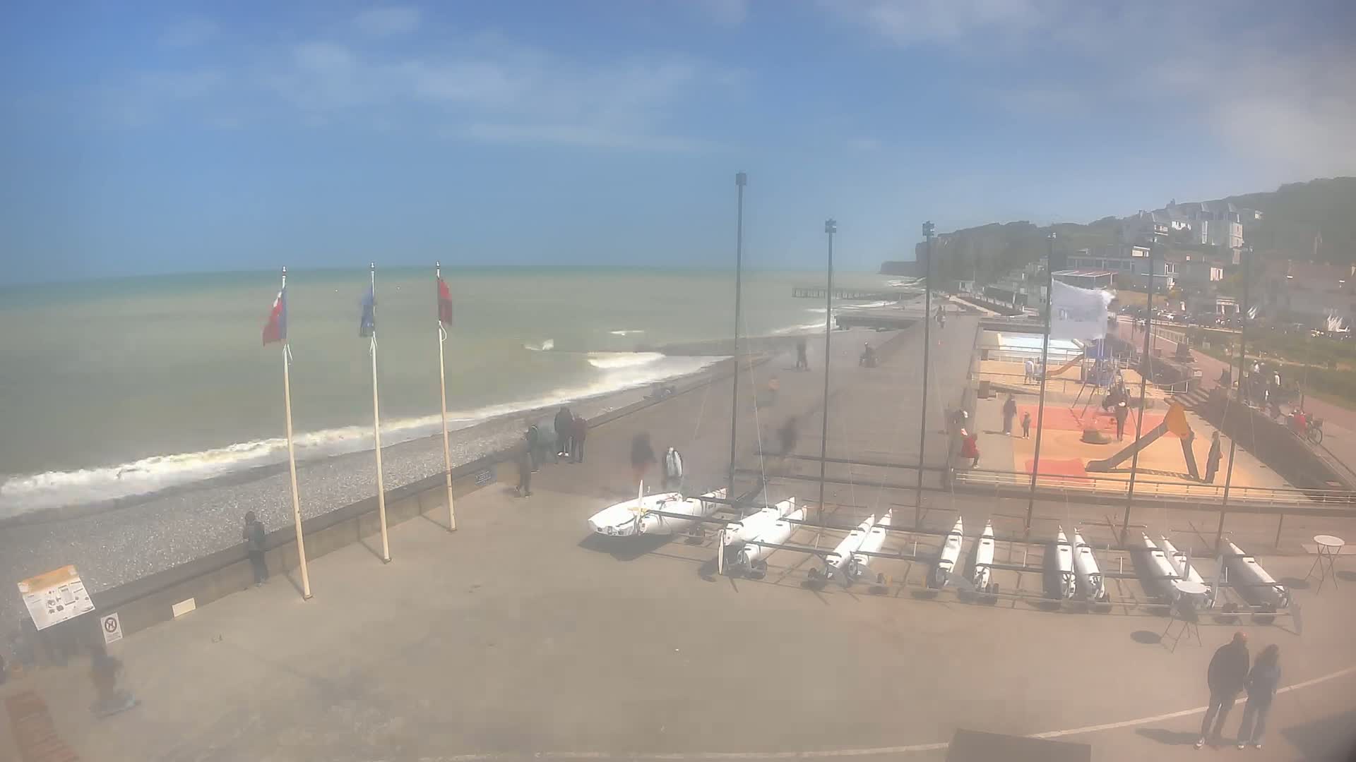 A mostly sunny day at a beach shows a row of small sailboats stored on a paved area next to a pebble beach with a calm sea, a playground, and several people scattered around.