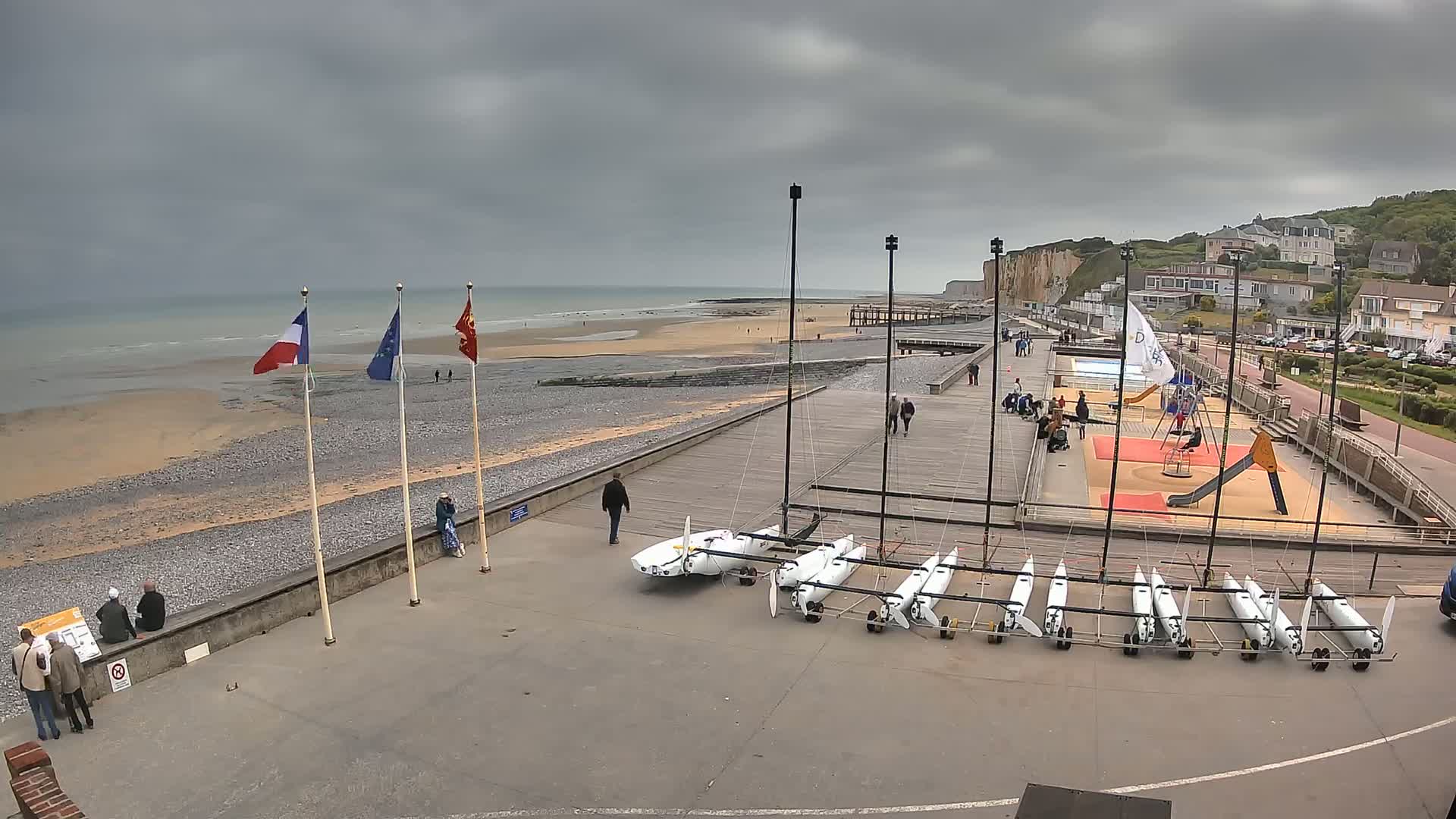 A beach with a gray overcast sky, several sailboats parked on a paved area next to a boardwalk, and a playground is visible near a residential area overlooking the ocean.