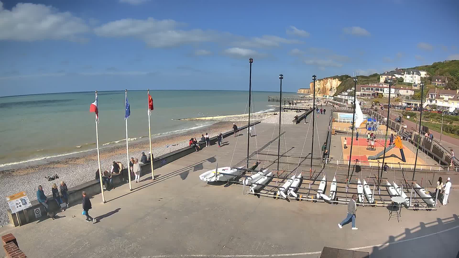 A sunny day at a seaside promenade shows several people gathered near a beach with white cliffs in the background, and a collection of small sailboats are lined up on the paved area.