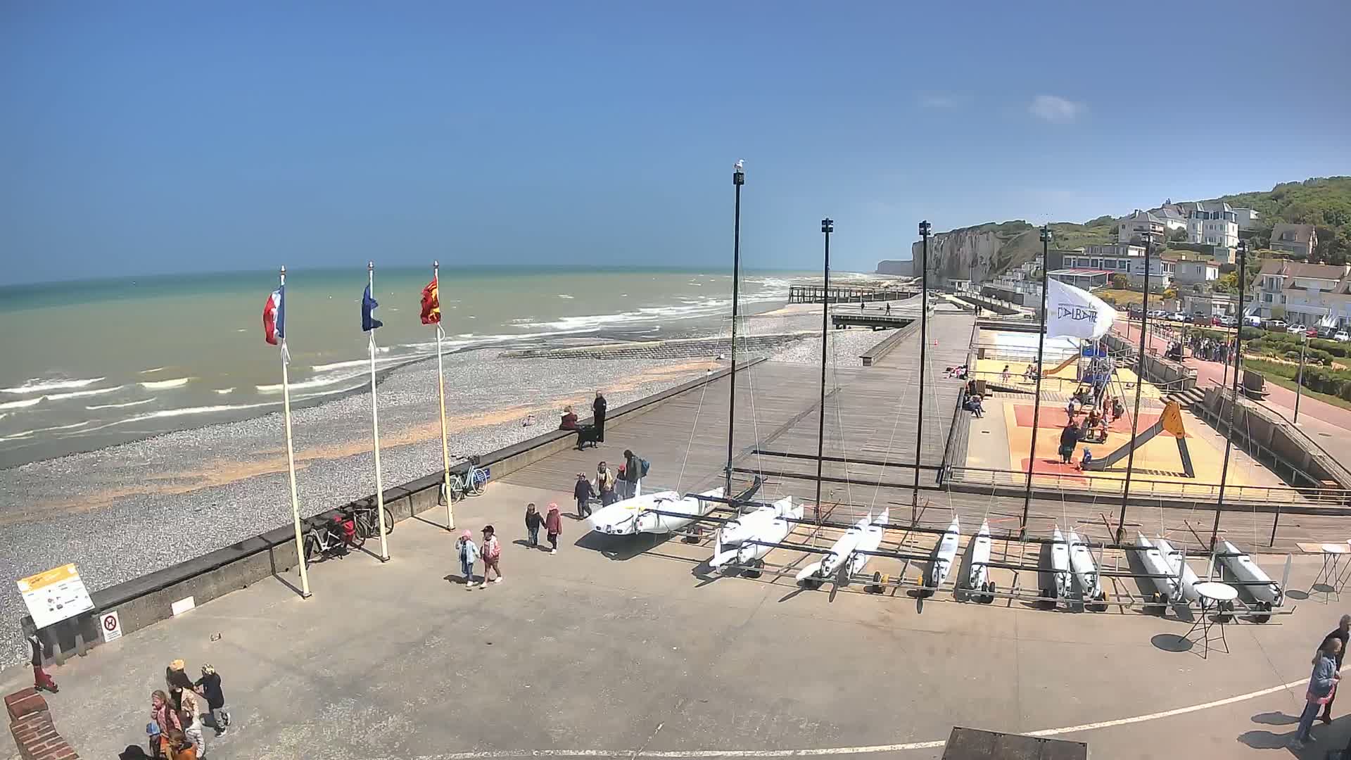 A sunny day at a seaside boardwalk shows several small sailboats stored near a playground and beach with a rocky shore and choppy waves.