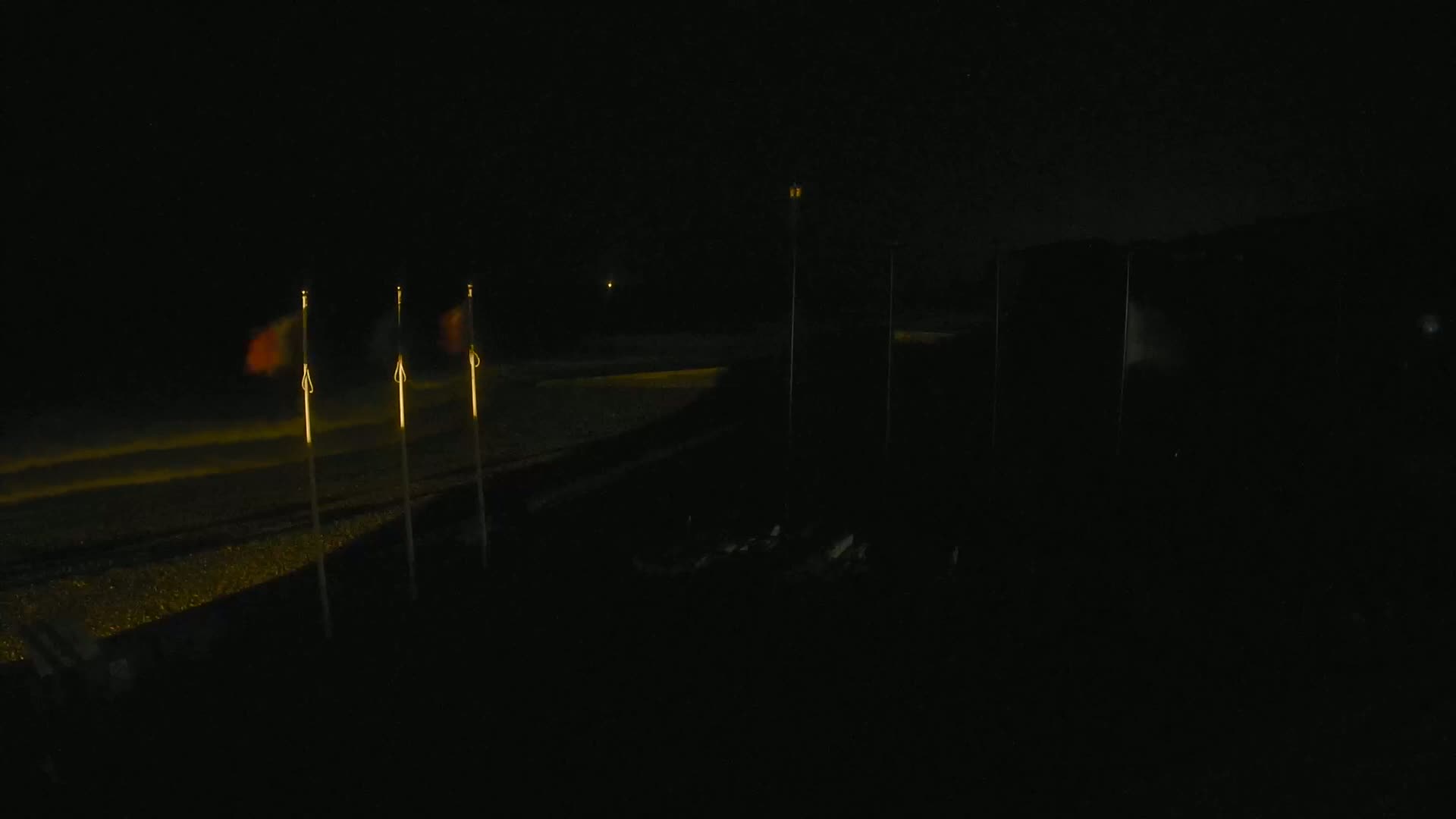 Several flags are illuminated on poles at night near a dimly lit area.