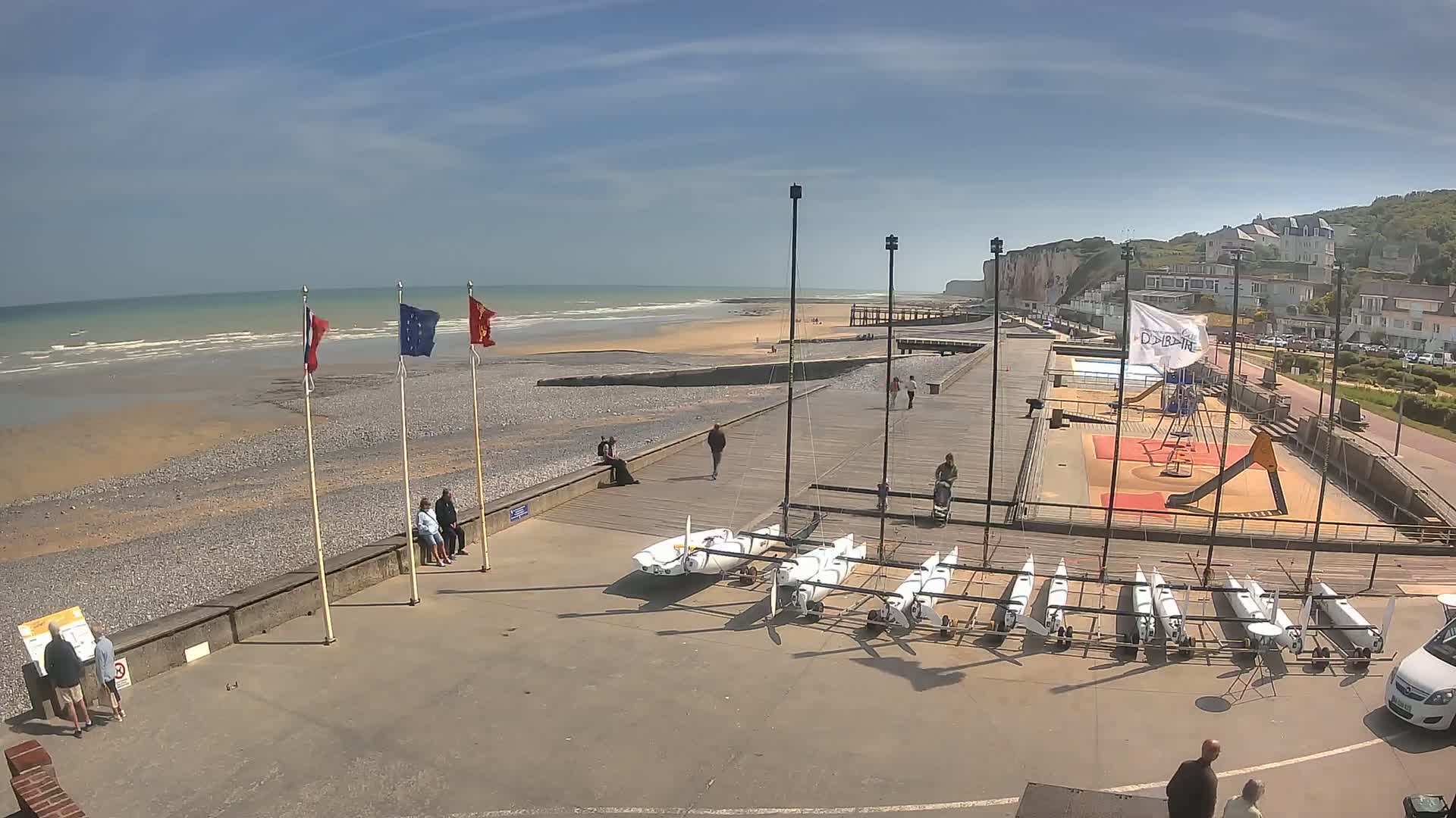 A beach with a pebble shore, several small sailboats, a playground, and a few people walking on a sunny day.