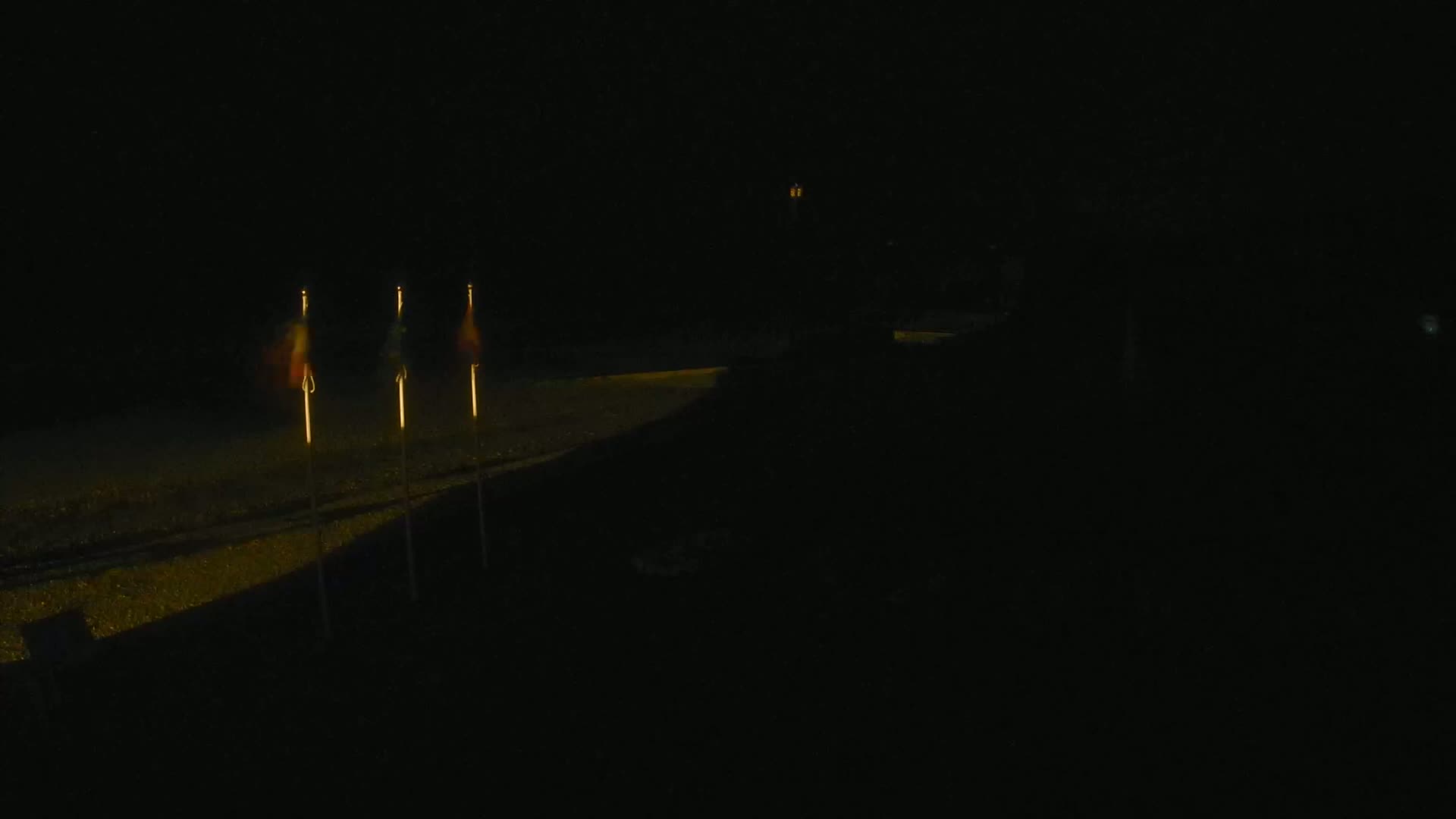 Three small flags are illuminated on tall poles at night next to a dark, paved area.