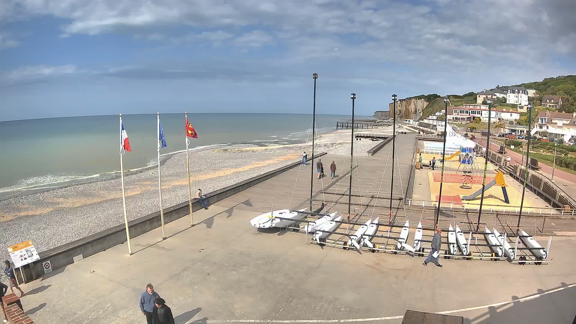 A mostly sunny day at a beach with several small sailboats docked on a paved area next to a pebble beach and a boardwalk, with white cliffs and buildings visible in the background.