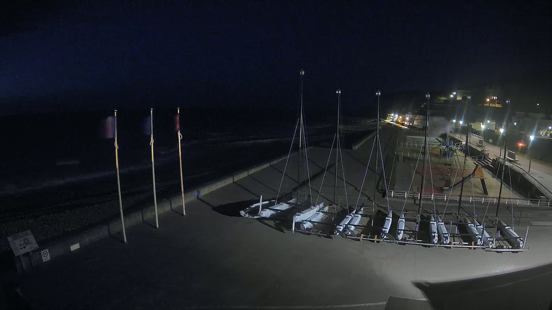 At night, several small sailboats are docked on a concrete surface next to a dark, wavy ocean under a clear, dark sky.