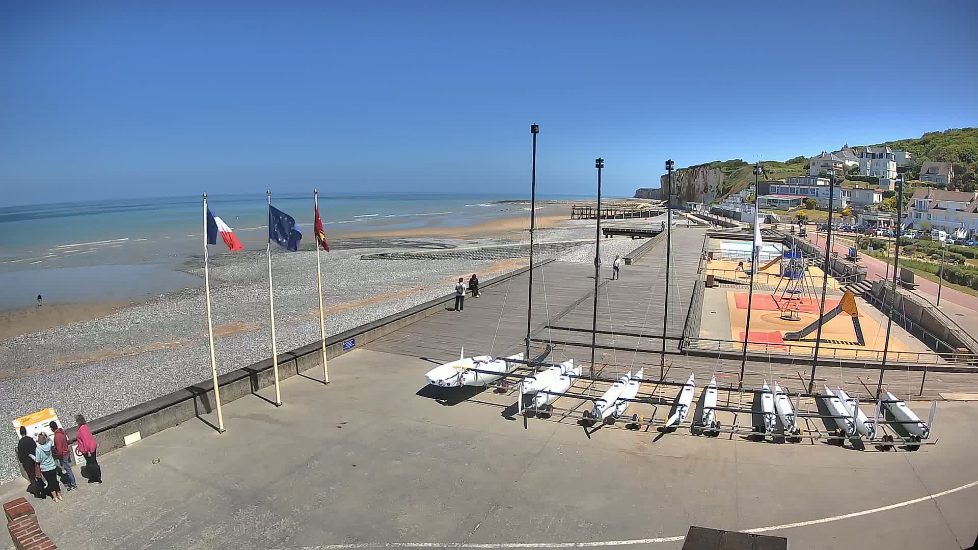 A pebble beach with a boardwalk, several sailboats, a playground, and French and European Union flags under a clear blue sky.