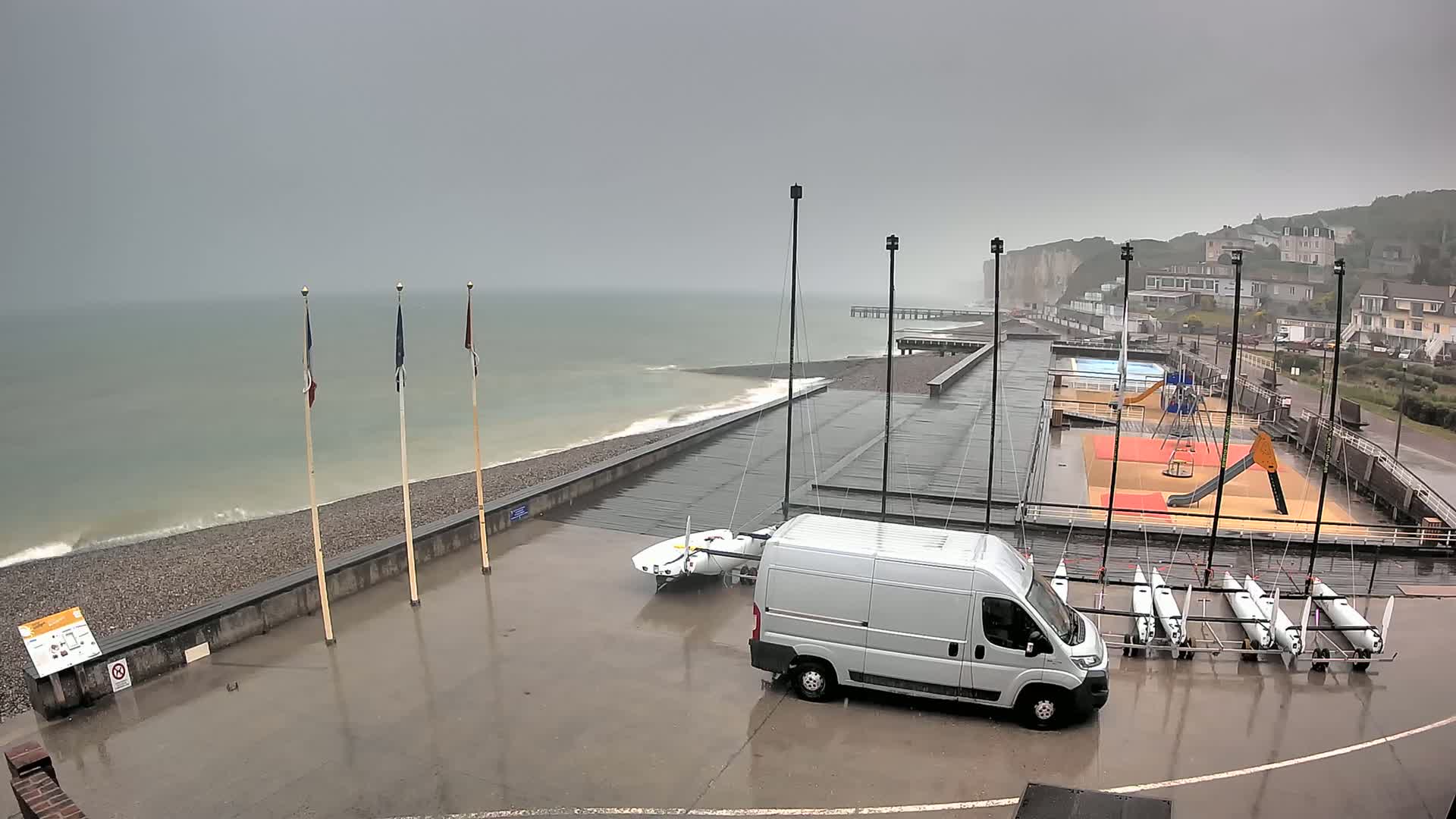 A rain-soaked coastal area features a white van parked near several sailboats, a playground, and a pier extending into a gray sea with small waves.