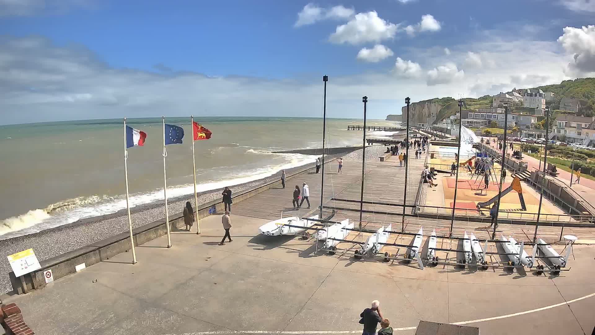 A seaside boardwalk with several small sailboats, a playground, and people walking, under a partly cloudy sky with some waves crashing on a pebbled beach.