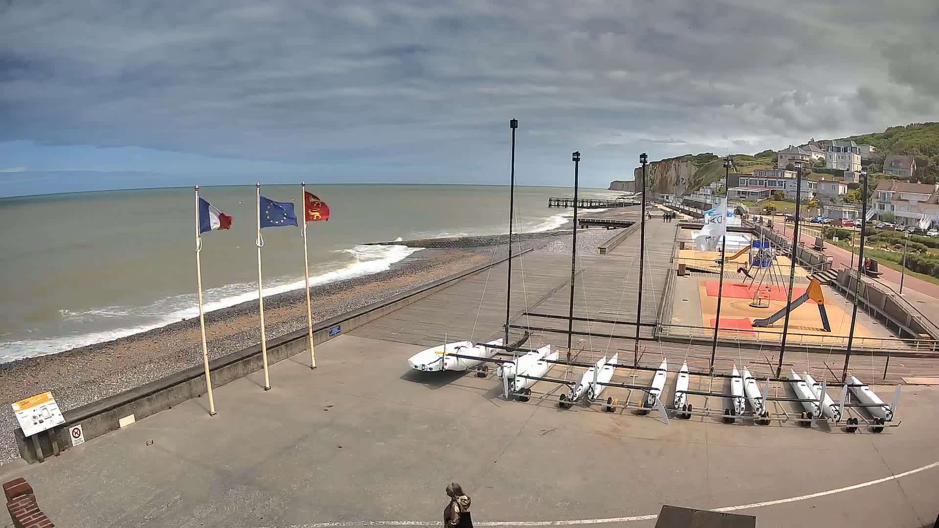 A seaside area with several small sailboats parked on a paved surface, a playground, a pier, and a rocky beach with small waves under a cloudy sky.