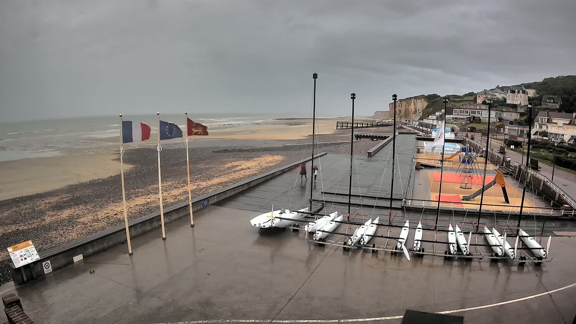 A wet, paved area next to a beach holds several small sailboats under a gray, overcast sky.