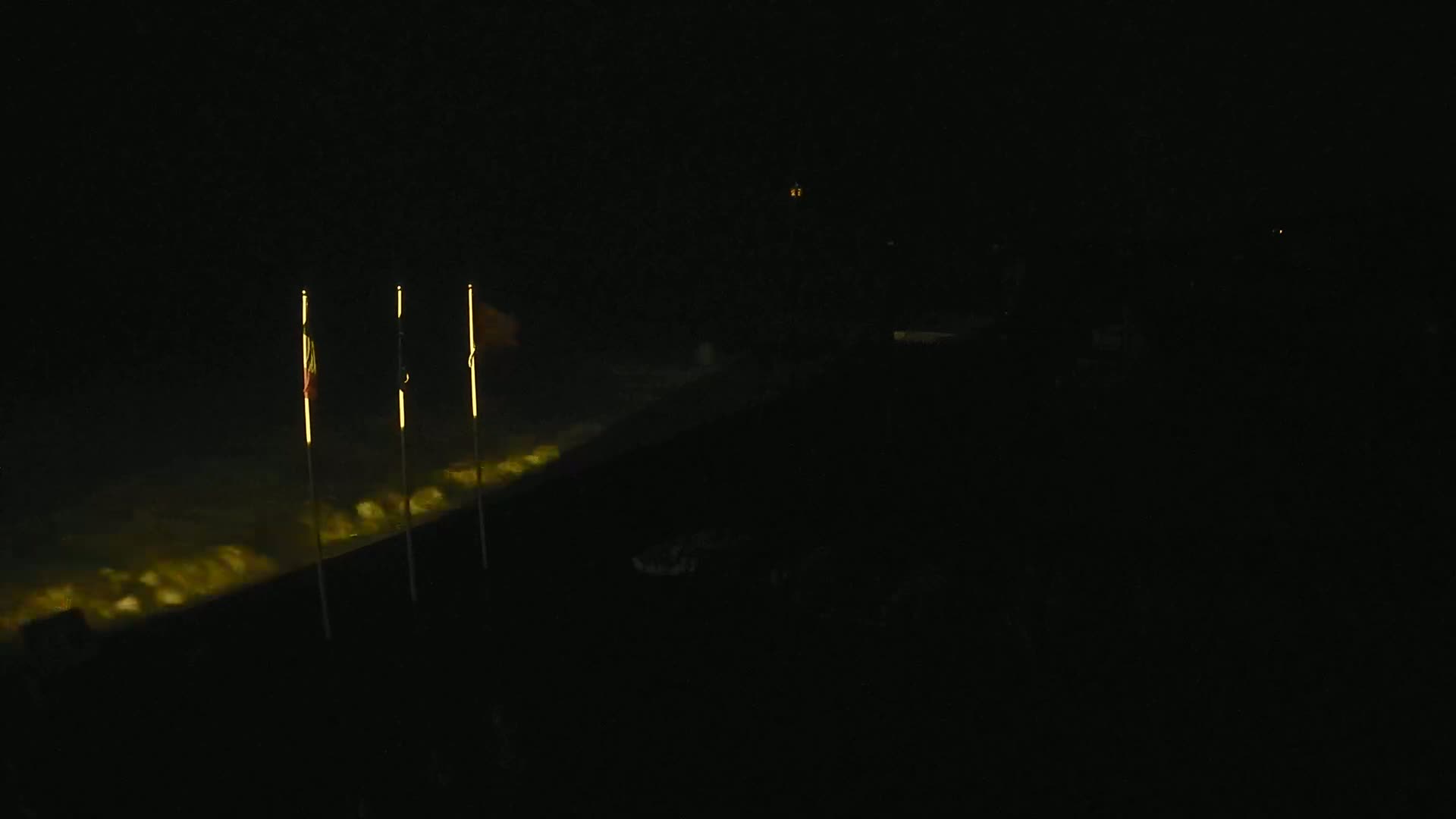 Three flags are illuminated on poles at night next to a dimly lit body of water.