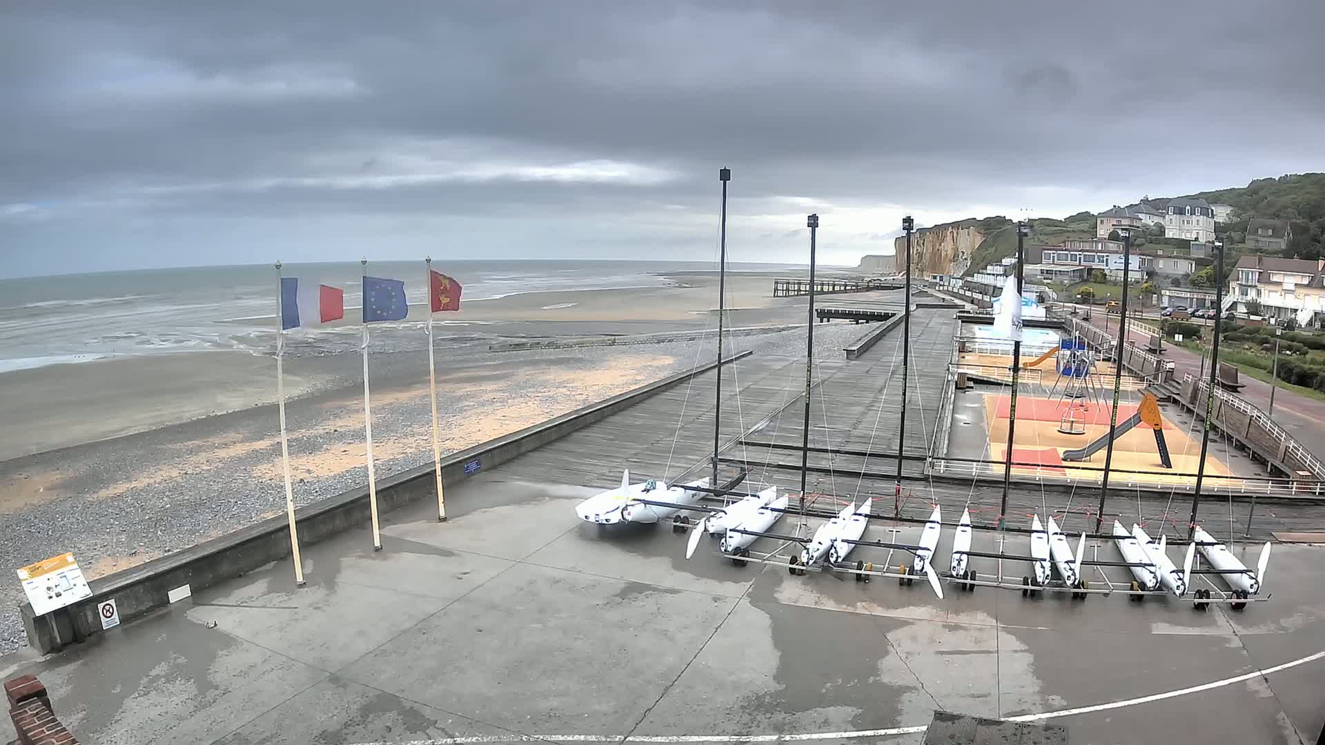 Several small sailboats are stored on a concrete area next to a beach with a wooden boardwalk, under an overcast sky.
