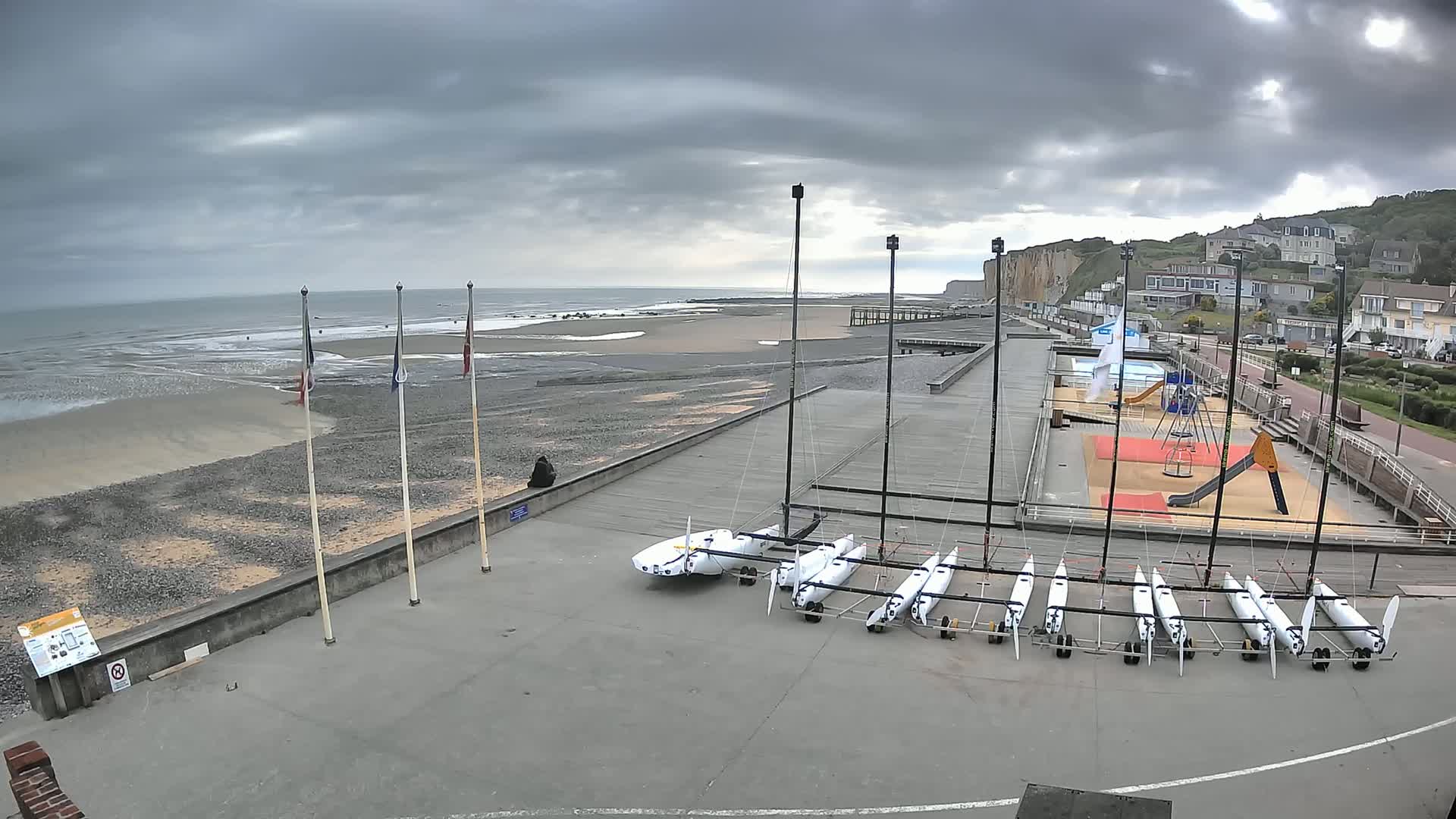 A beach with several sailboats parked on a paved area next to a playground, under a cloudy sky.