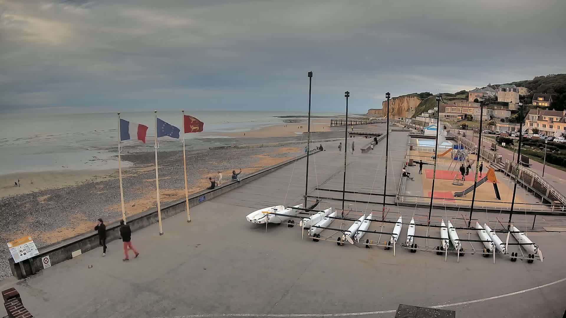 A beachside promenade with several small sailboats, a playground, and a few people walking under an overcast sky.