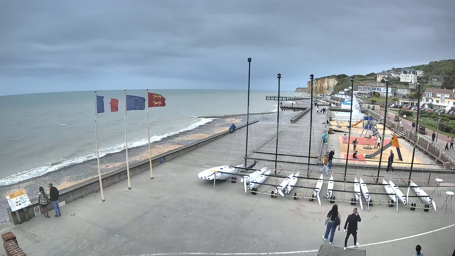A mostly cloudy day at a seaside location shows several small sailboats docked on a paved area next to a beach with a rocky shore, a playground, and a boardwalk leading toward white cliffs and buildings in the distance.