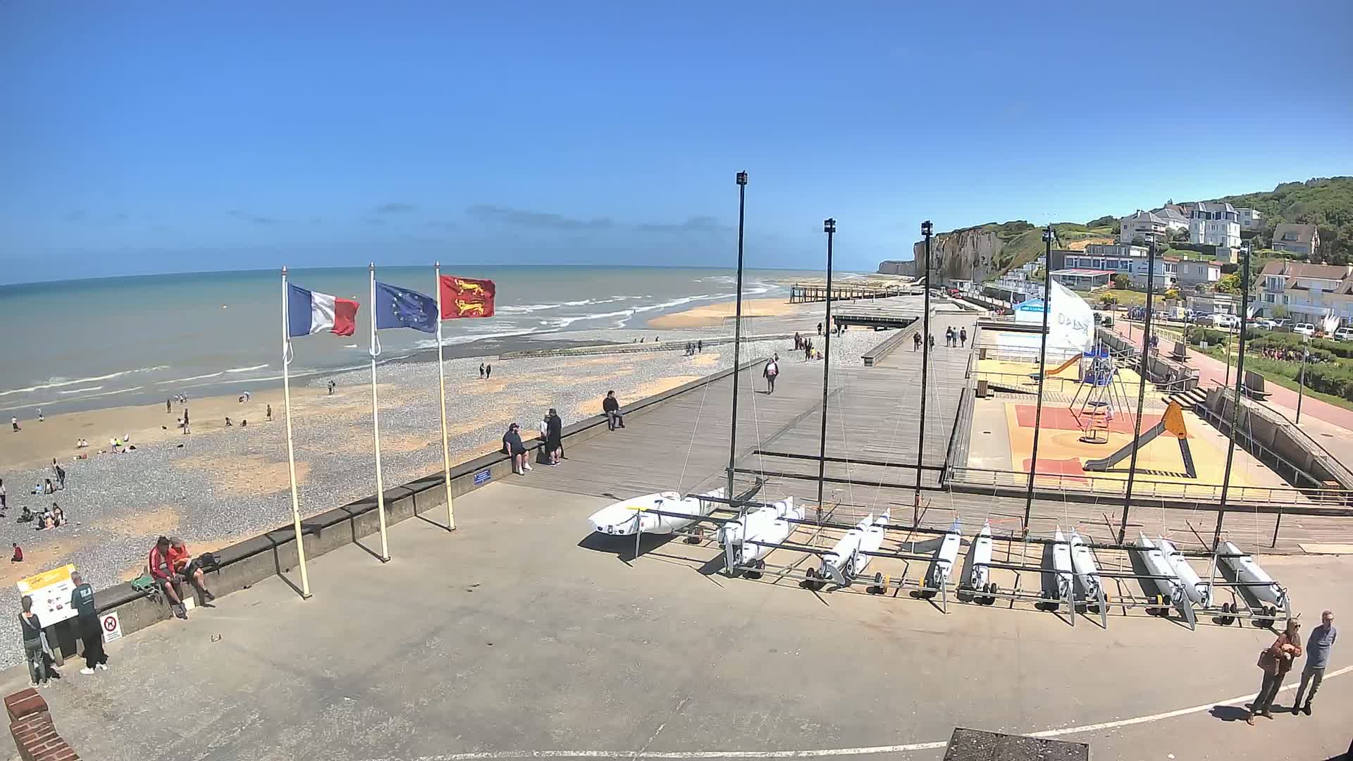 A sunny day at a beach with several people relaxing, a playground, and small sailboats stored on the shore next to a boardwalk.