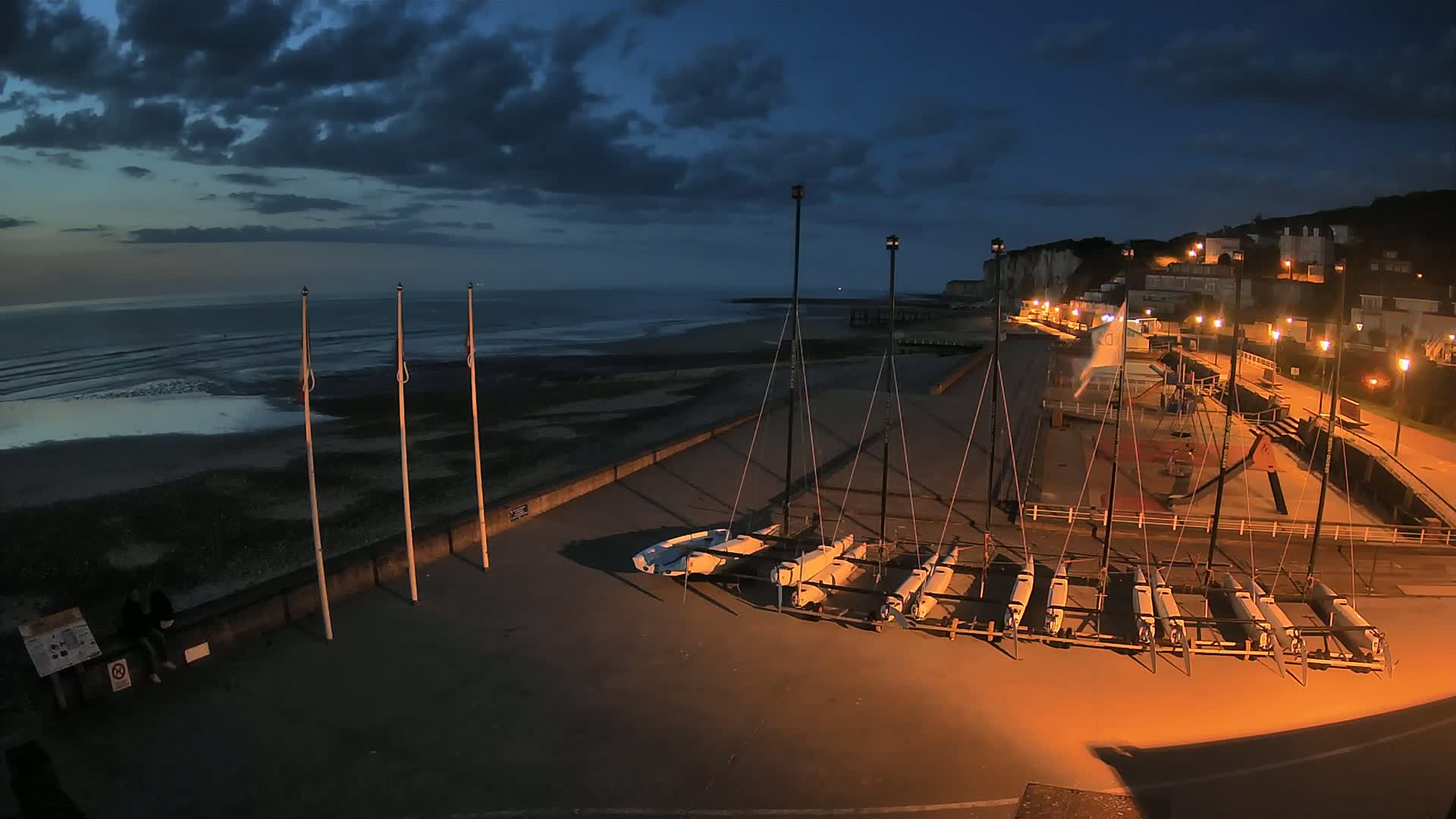 Several sailboats are docked on a paved area next to a beach at twilight under a dark, cloudy sky.