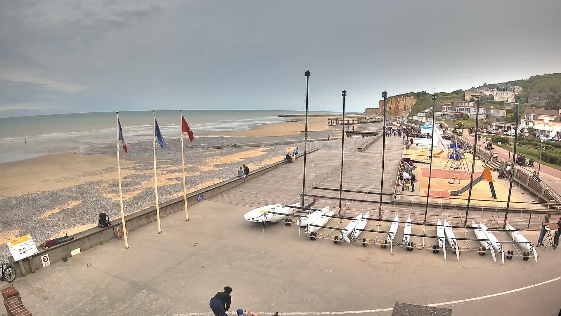 A beach with a mostly cloudy sky, several small sailboats, a playground, and people scattered along a paved walkway and beach.