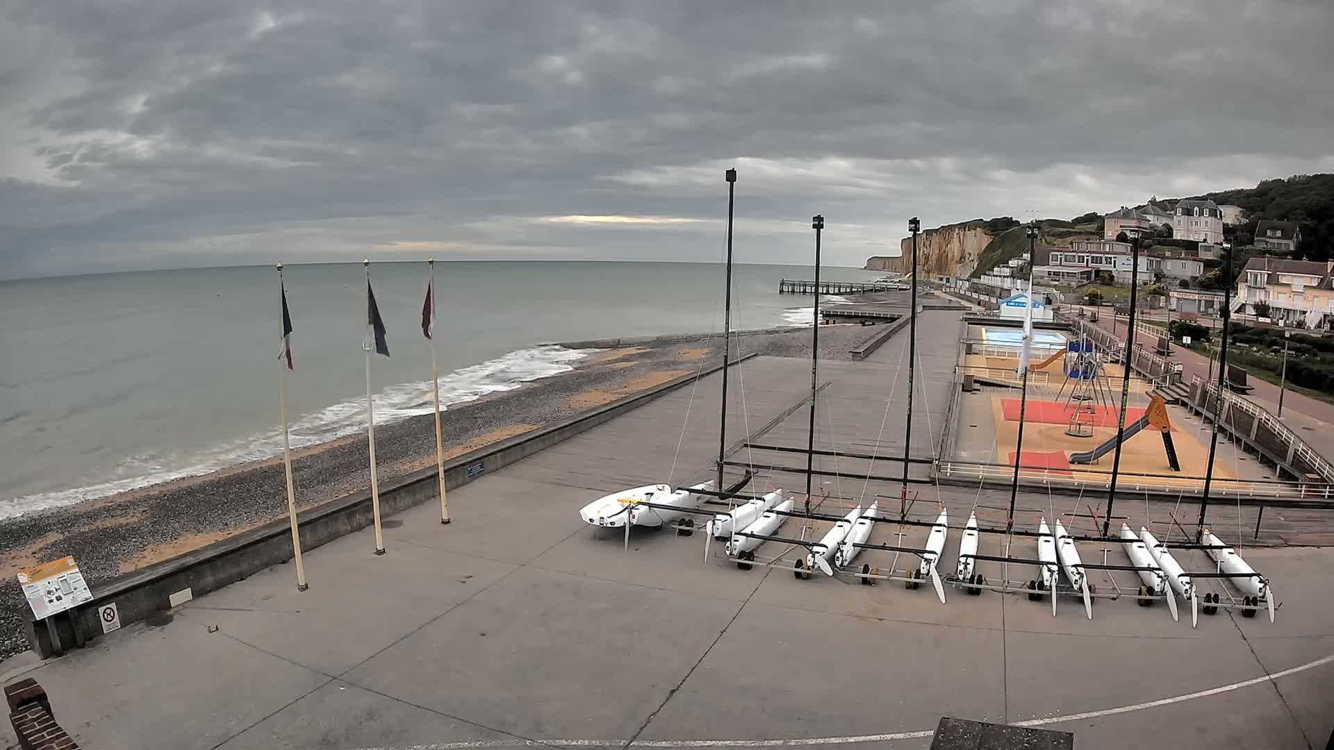 A pebble beach with several sailboats stored on a paved area next to a playground, under an overcast sky, overlooks a calm sea and white cliffs.