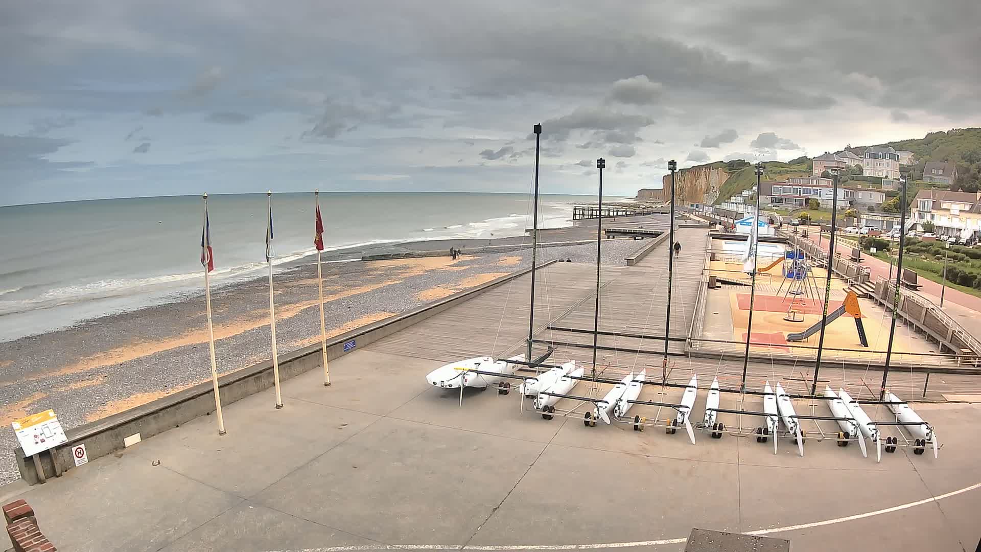 A beach with several small sailboats stored on a paved area next to a playground, overlooking a calm sea under a cloudy sky.