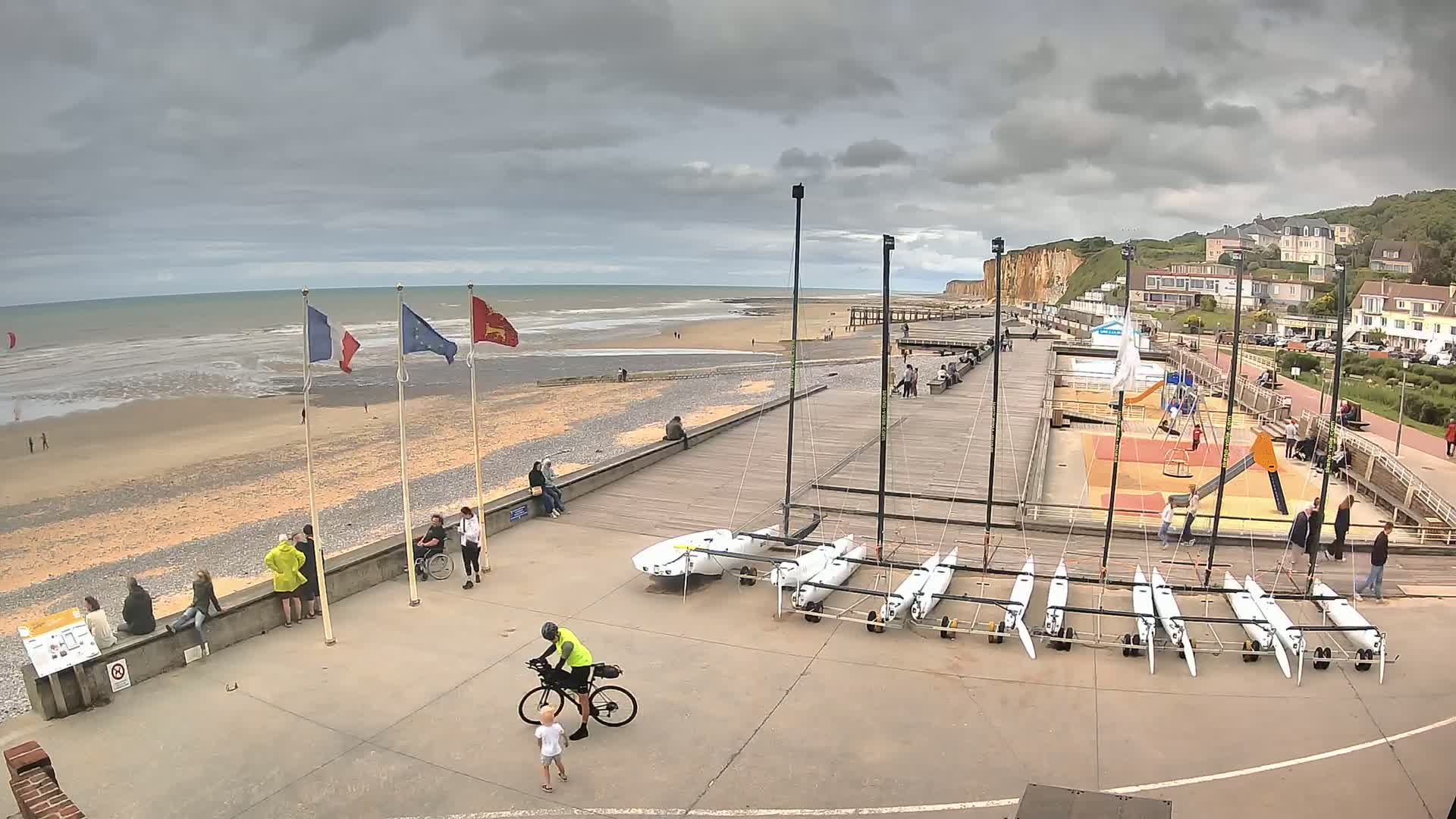 A mostly cloudy day at a beach with several people scattered around a boardwalk area near white sailboats, a playground, and white cliffs in the distance.