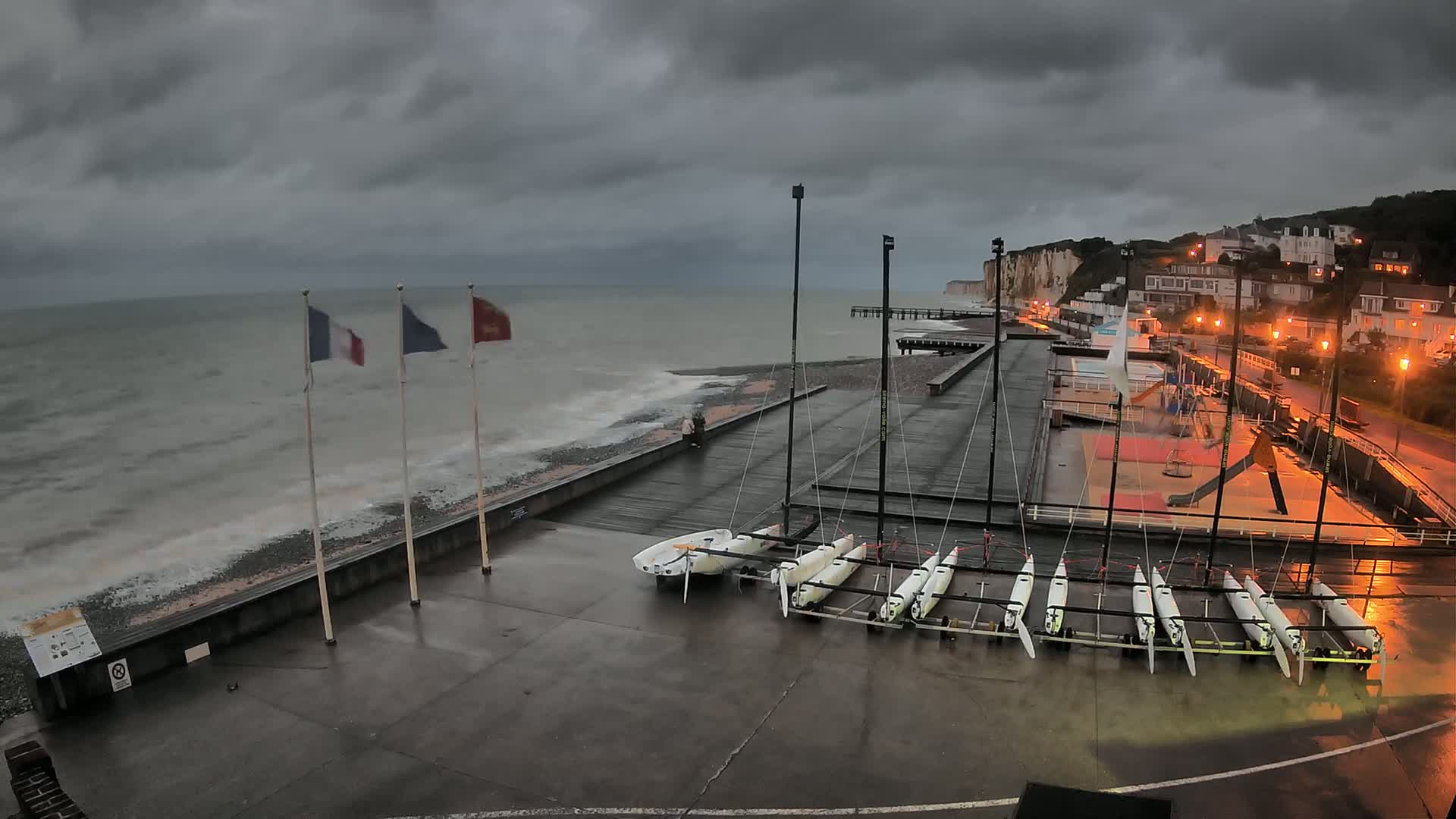 Several small sailboats are docked on a wet concrete area next to a rocky beach with choppy gray water under a dark, cloudy sky.