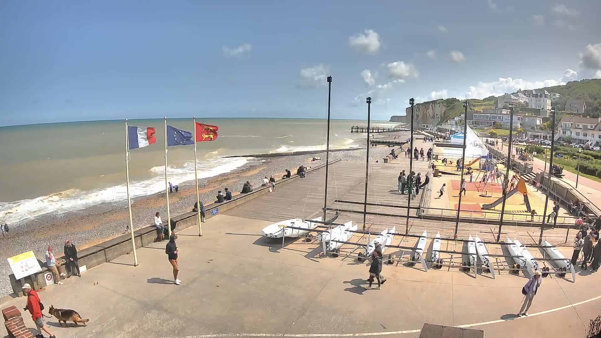 A partly sunny day at a beachside boardwalk shows people relaxing, children playing, and small sailboats stored near a rocky shoreline and white cliffs.