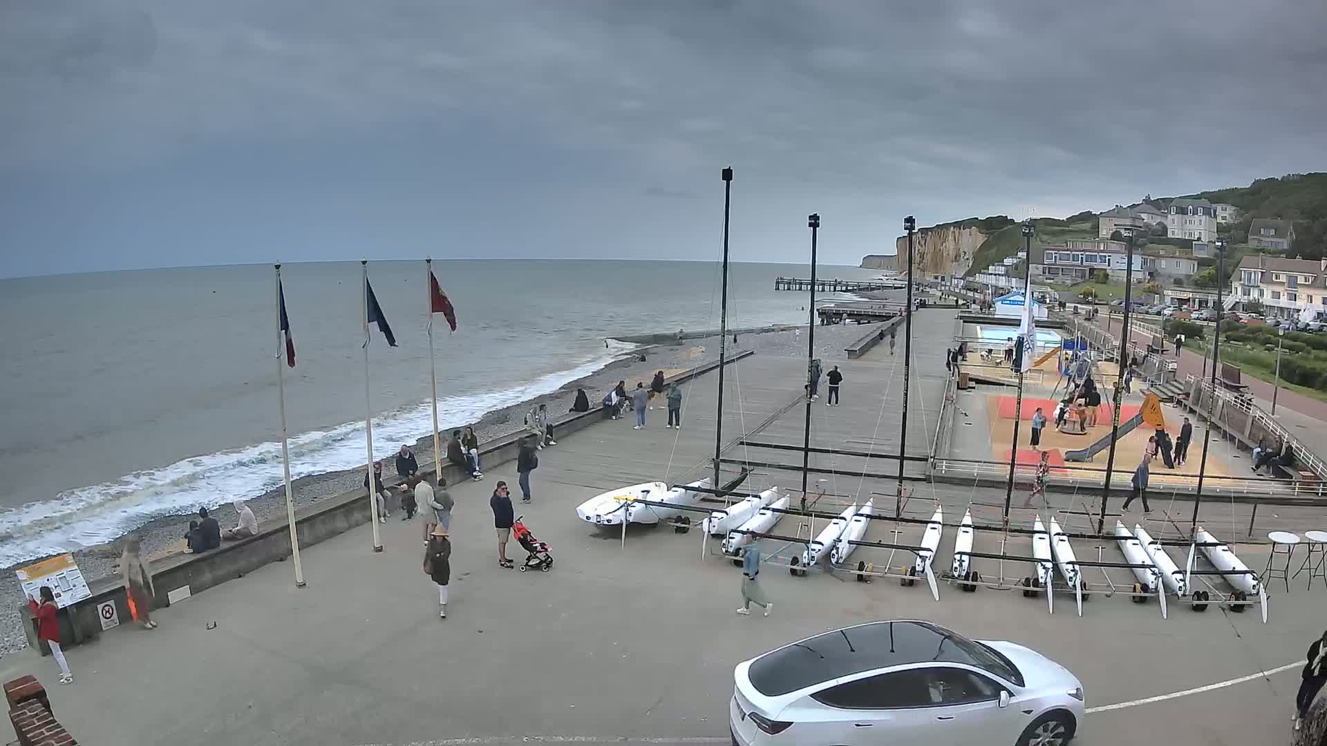 A seaside town with overcast skies shows people gathered near a beach, several small sailboats, a playground, and a white car parked near the water.