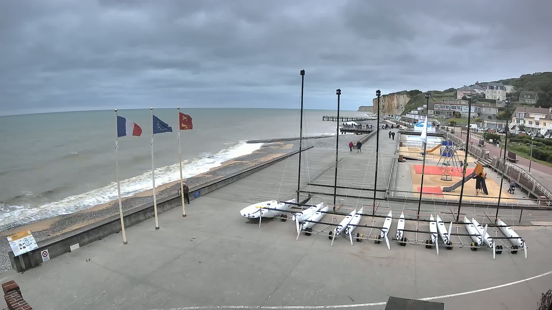 A seaside promenade with several small sailboats, a playground, and a pier under an overcast sky, with gentle waves breaking on a pebble beach.