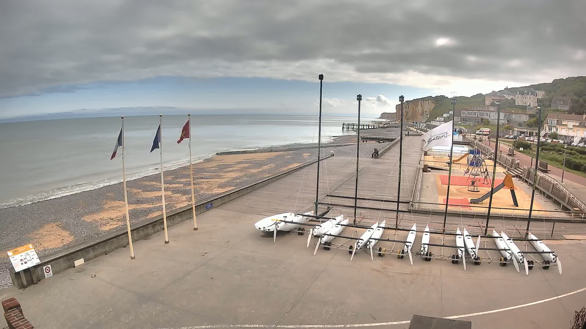 A mostly cloudy day at a seaside location shows several small sailboats stored on a paved area next to a beach, a playground, and a pier extending into the ocean.