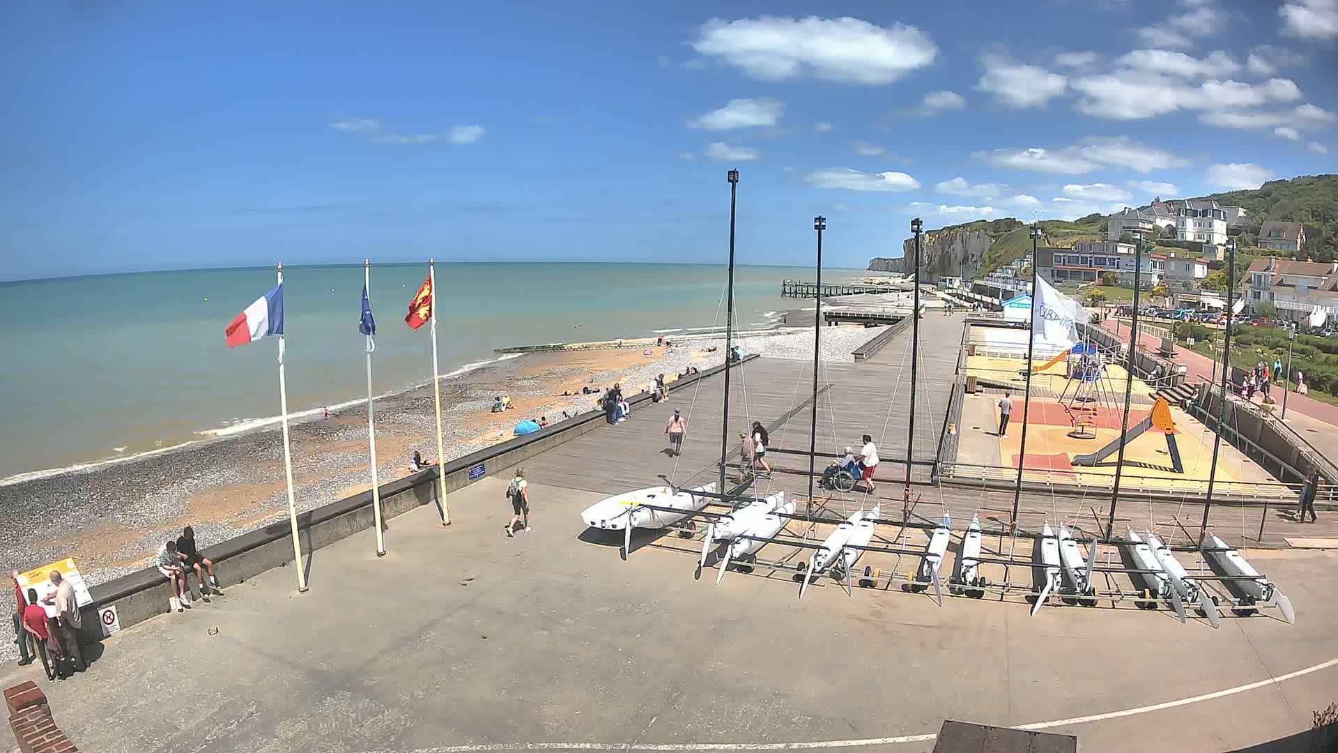 A sunny day at a seaside town shows a beach with people, several small sailboats on trailers, a playground, and a walkway along white cliffs.