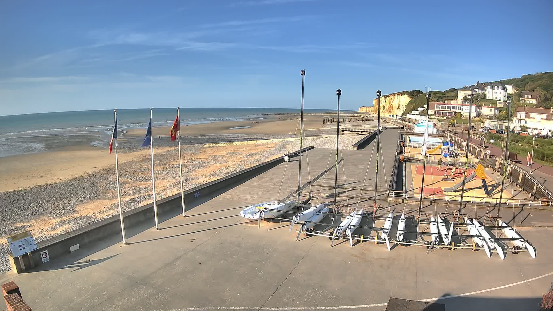 A sandy beach with several sailboats docked near a paved area with a playground, backed by white cliffs and a residential area under a clear blue sky.