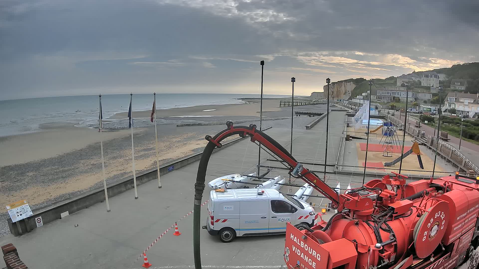 A red vacuum truck is parked near a beach with overcast skies, a white van, and a playground visible in the background.