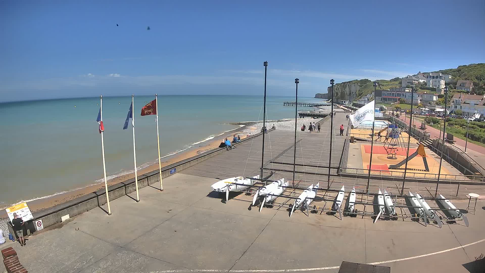 A sunny day at a seaside boardwalk shows several small sailboats stored on the ground near a playground, a pier extending into a calm sea, and white cliffs in the background.