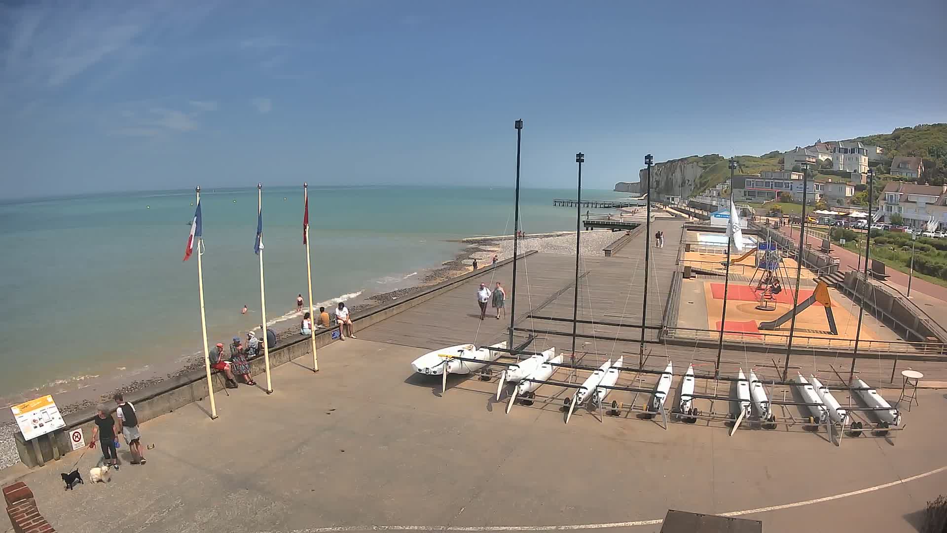 A sunny seaside scene shows a boardwalk, several small sailboats, a playground, and people relaxing along a rocky beach with white cliffs in the distance.