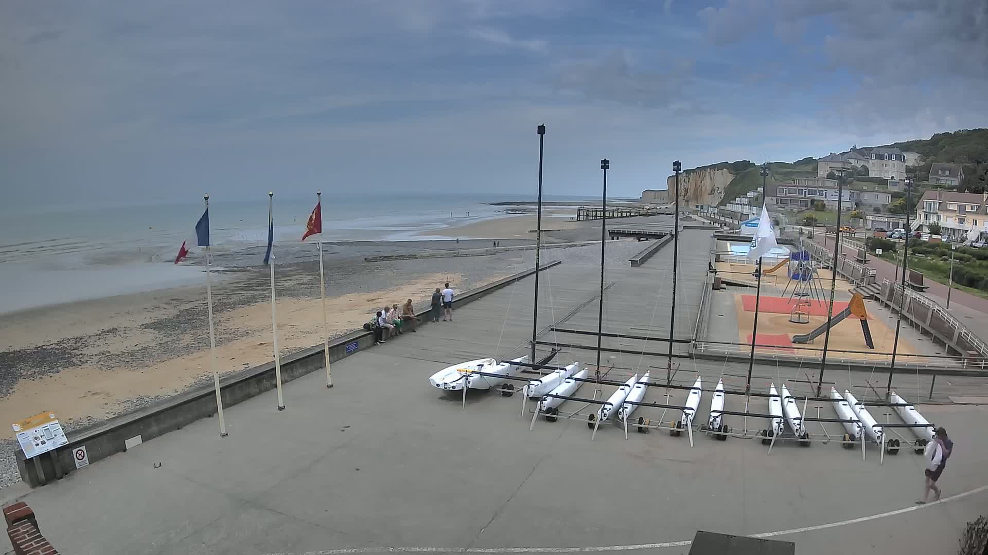 A mostly cloudy day shows a beach with several small sailboats stored on a paved area near a playground, a seawall, and white cliffs in the distance.