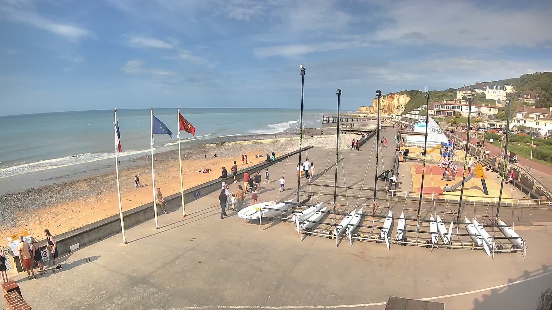 A beach scene on a sunny day shows people relaxing and playing near a row of small sailboats on a paved area, with white cliffs in the distance.