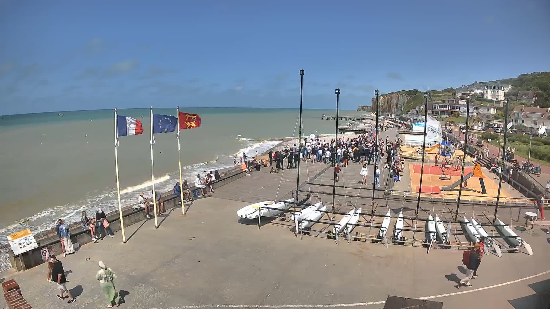 A sunny day at a seaside location shows a crowd of people gathered near the shore, several small sailboats lined up on a paved area, and a playground nearby.