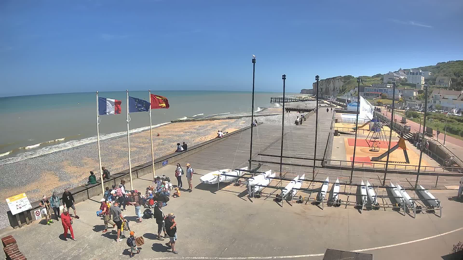 A pebble beach with a boardwalk, several small sailboats, a playground, and people gathered on a sunny day near white cliffs.