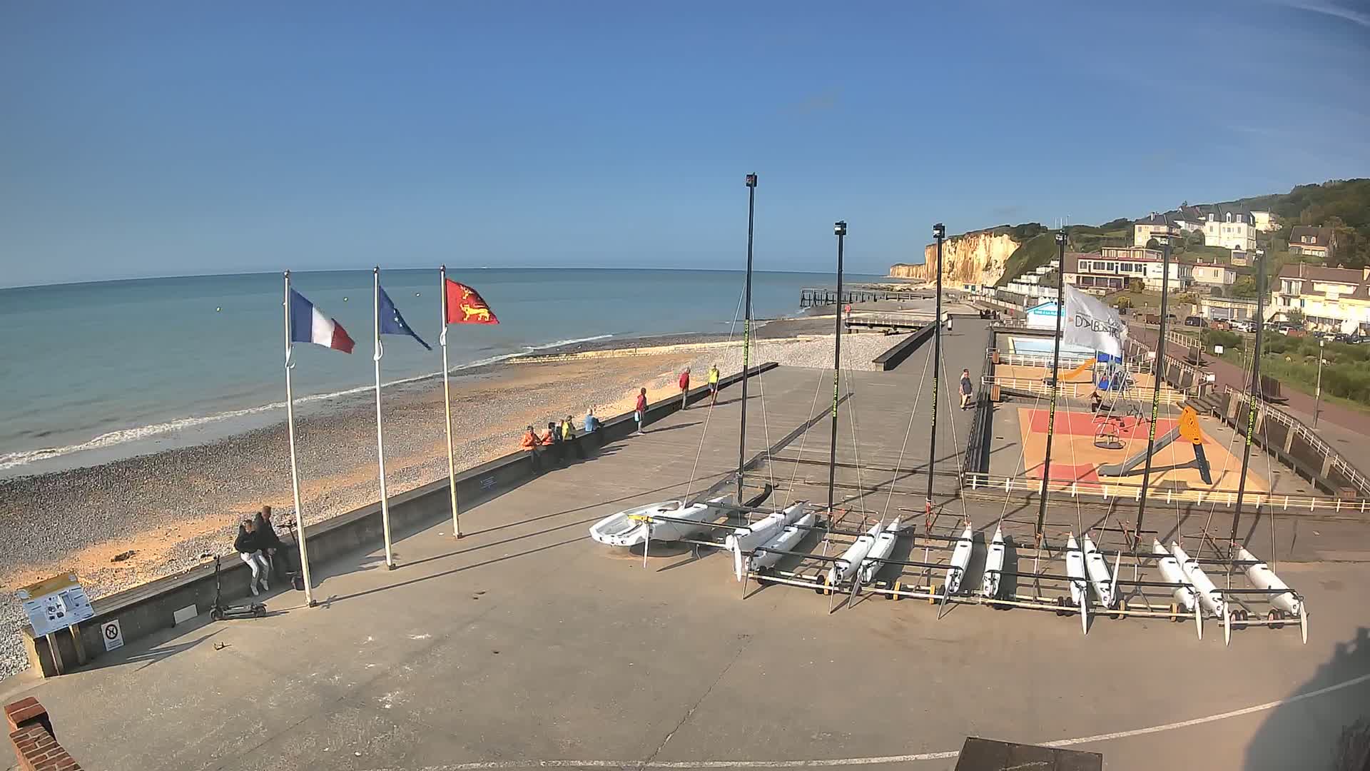 On a sunny day, several small sailboats are docked on a paved area near a beach with white cliffs in the background, and a few people are walking or sitting along the shore.