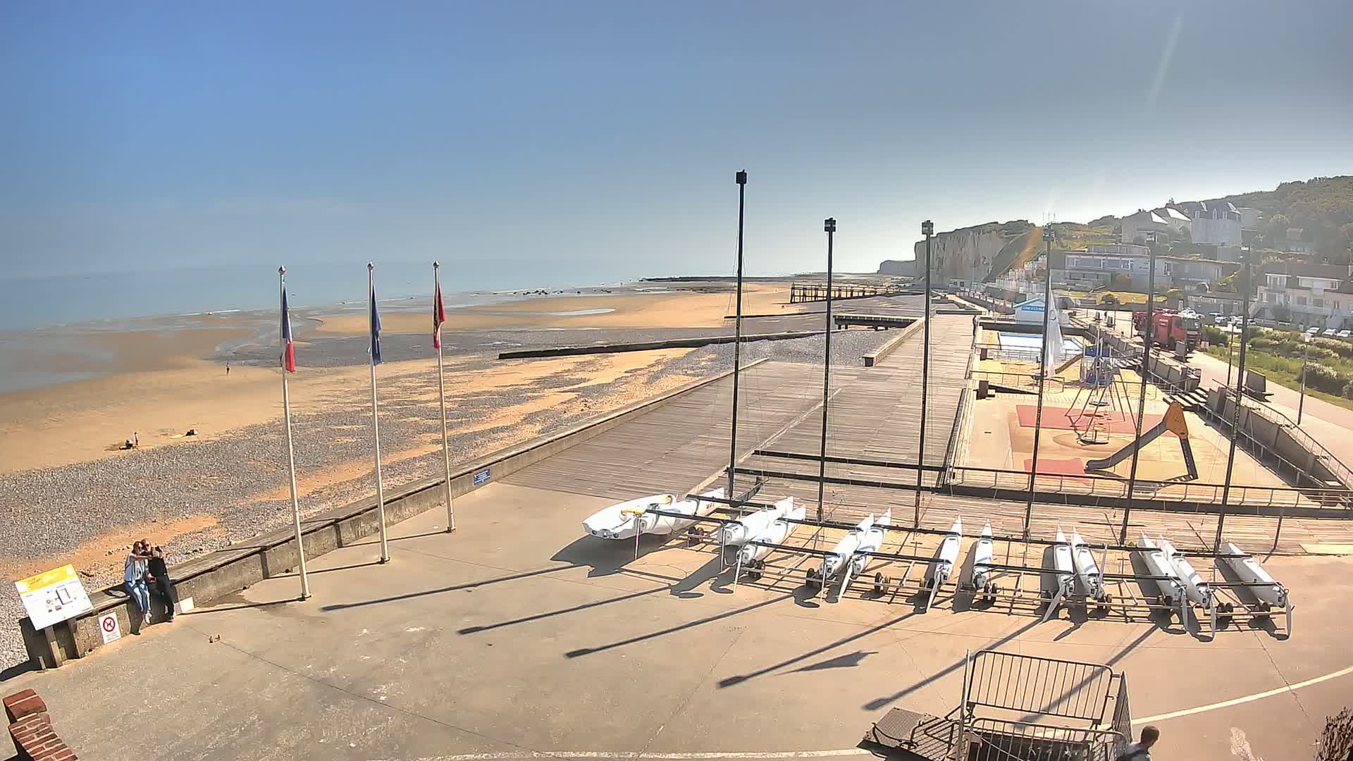 A sandy beach with a boardwalk and several small sailboats stored on racks, under a clear blue sky.
