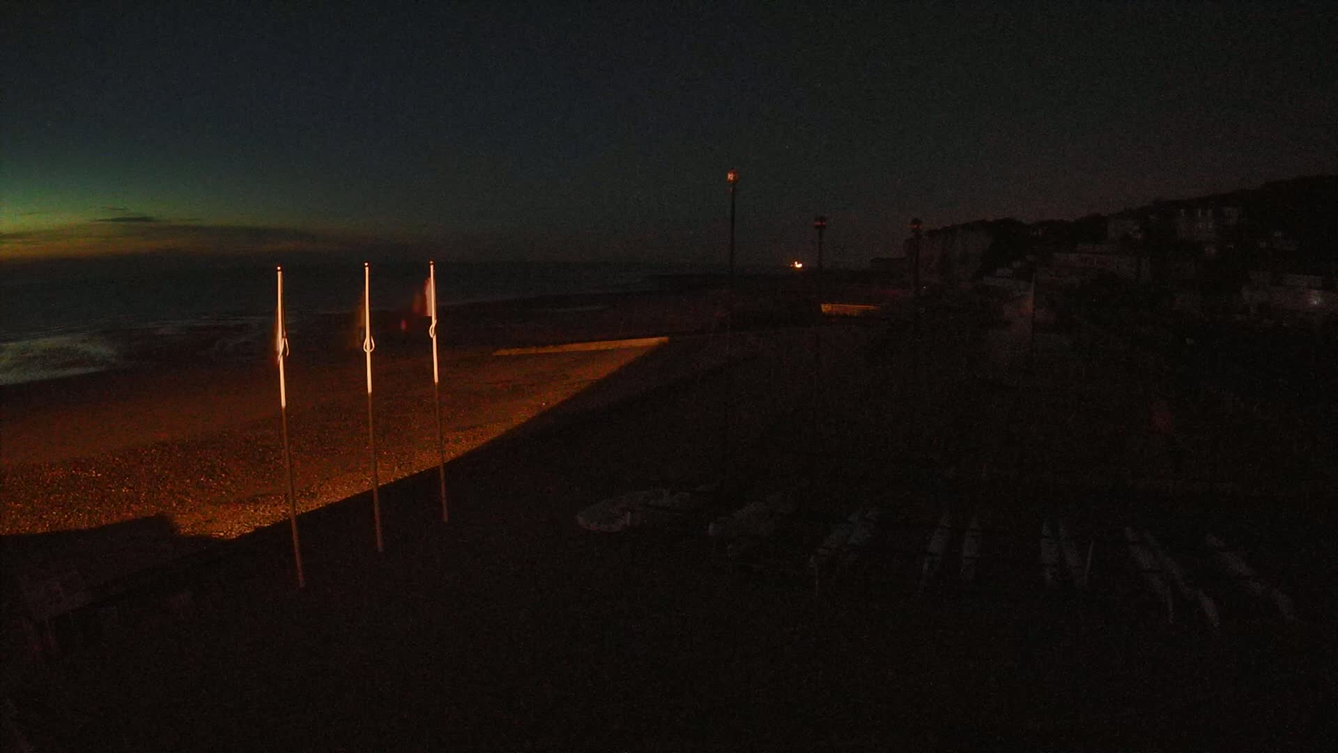 Three illuminated flagpoles stand on a dark beach at night near a dimly lit building under a mostly clear, twilight sky.