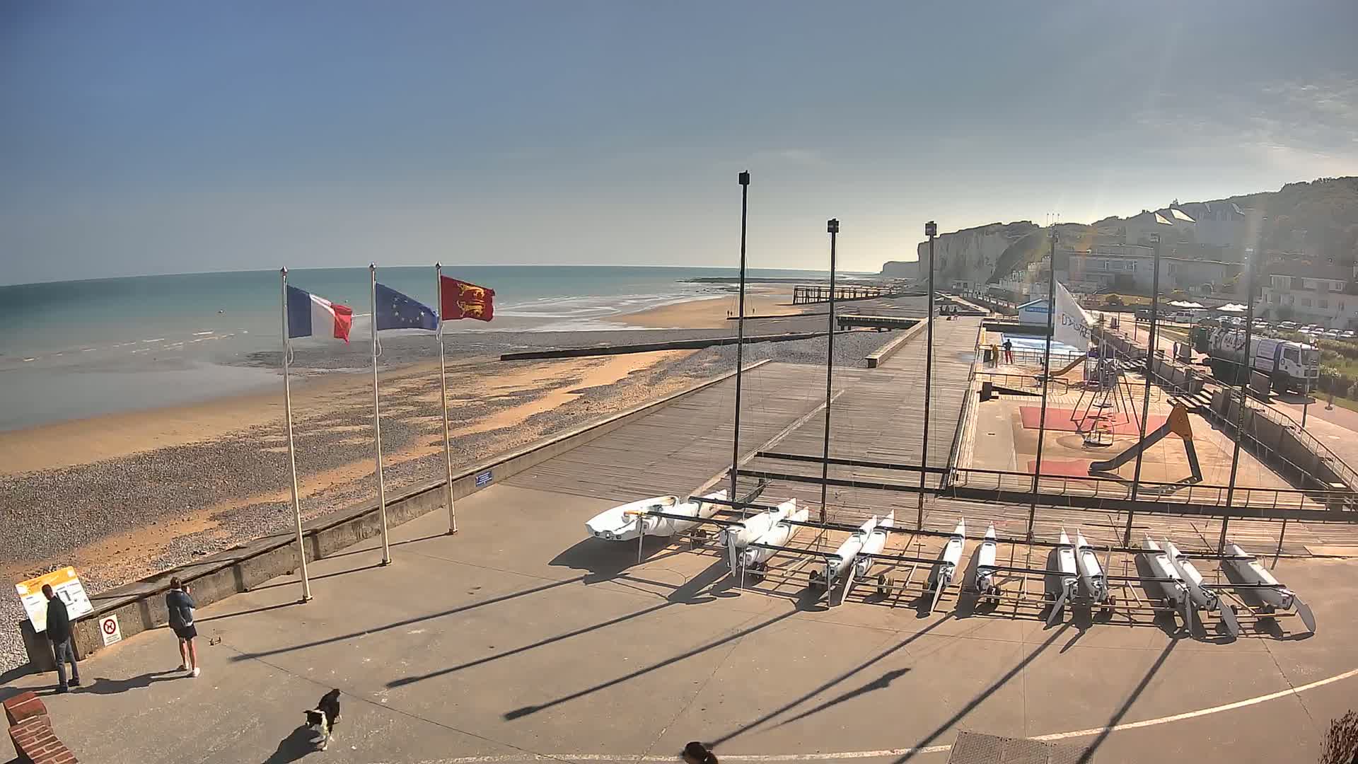 A beach with a boardwalk, several small sailboats, a playground, and white cliffs in the background under sunny skies.