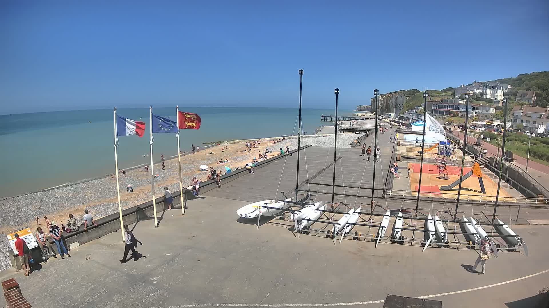 A sunny beach scene shows a pebbled shore with people sunbathing, several sailboats stored on the paved area near a boardwalk, and white cliffs in the background.