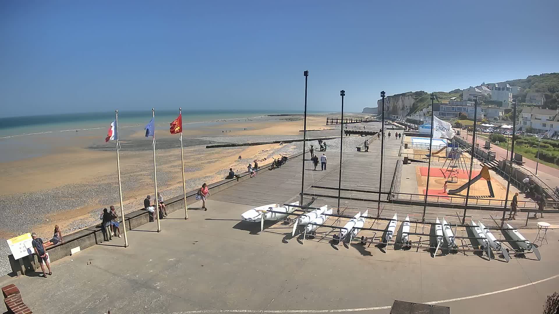 A sunny beach scene shows a boardwalk with people relaxing and several small sailboats lined up near a playground, all situated against a backdrop of white cliffs and calm ocean waters.