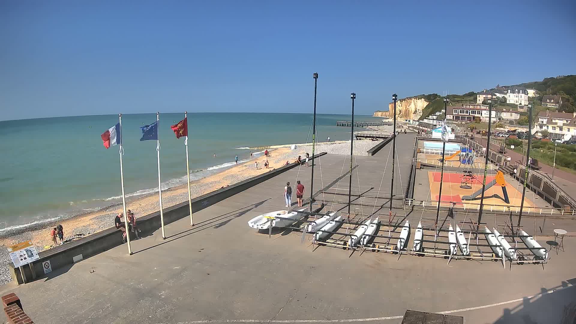 A sunny day at a beachside area shows several small sailboats docked near a playground and a calm ocean with white sand and a few people enjoying the beach.