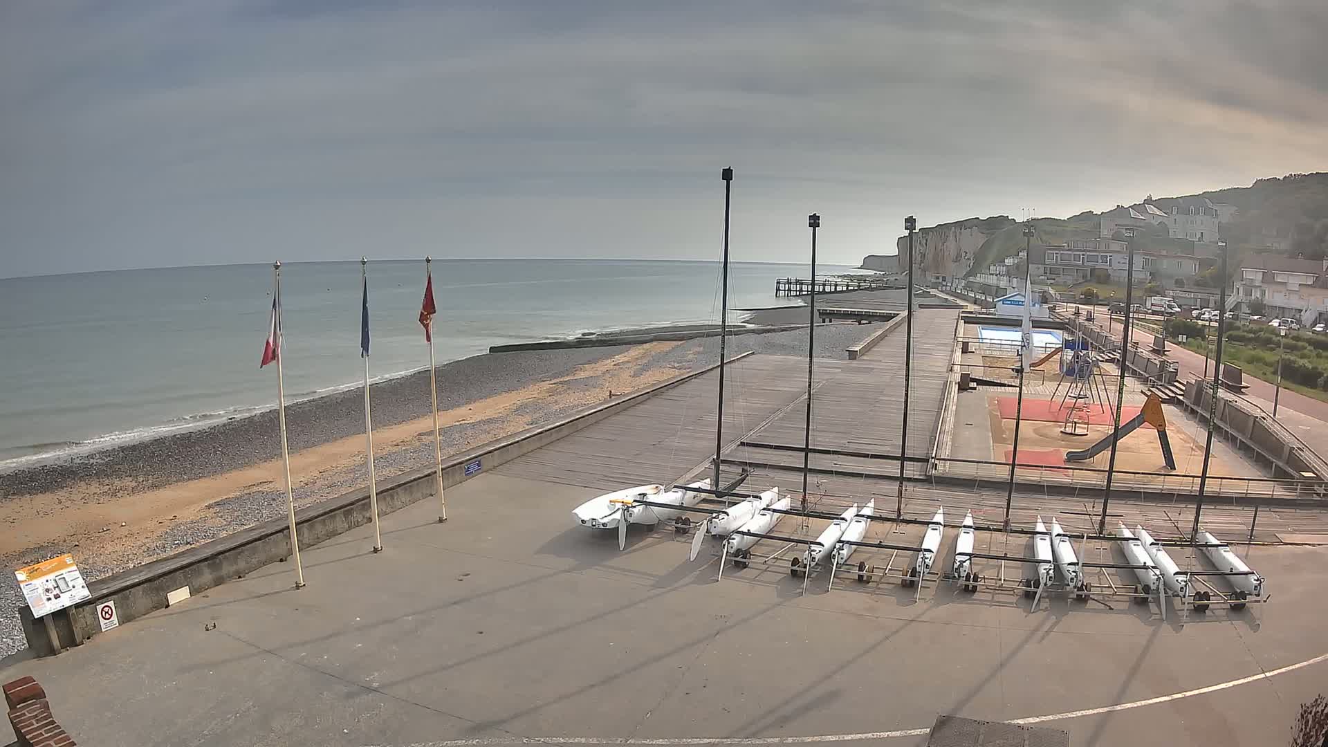 A beach with several small sailboats stored on trailers near a playground and a calm, gray sea under an overcast sky.