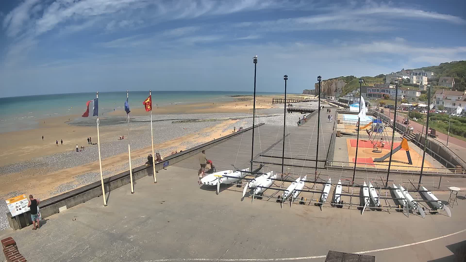 A sandy beach with people sunbathing and swimming, a boardwalk with several small sailboats parked, a playground, and white cliffs in the background under a partly cloudy sky.