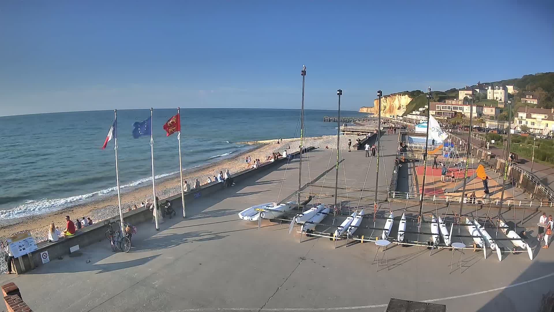 A beach with people relaxing, several sailboats stored on the paved area next to the water, and white cliffs in the background under a clear blue sky.