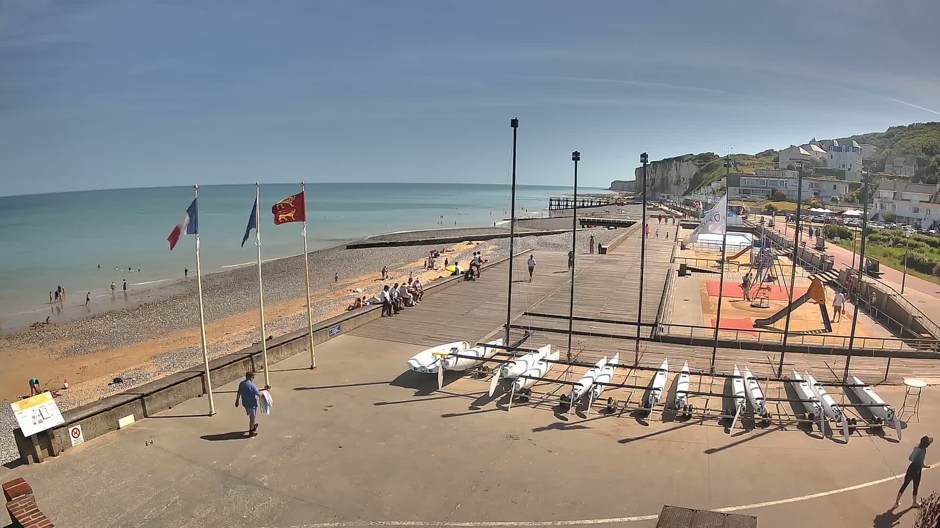 A beach scene on a sunny day shows people relaxing and swimming in the ocean, with several sailboats stored on a nearby paved area next to a playground and white cliffs in the background.